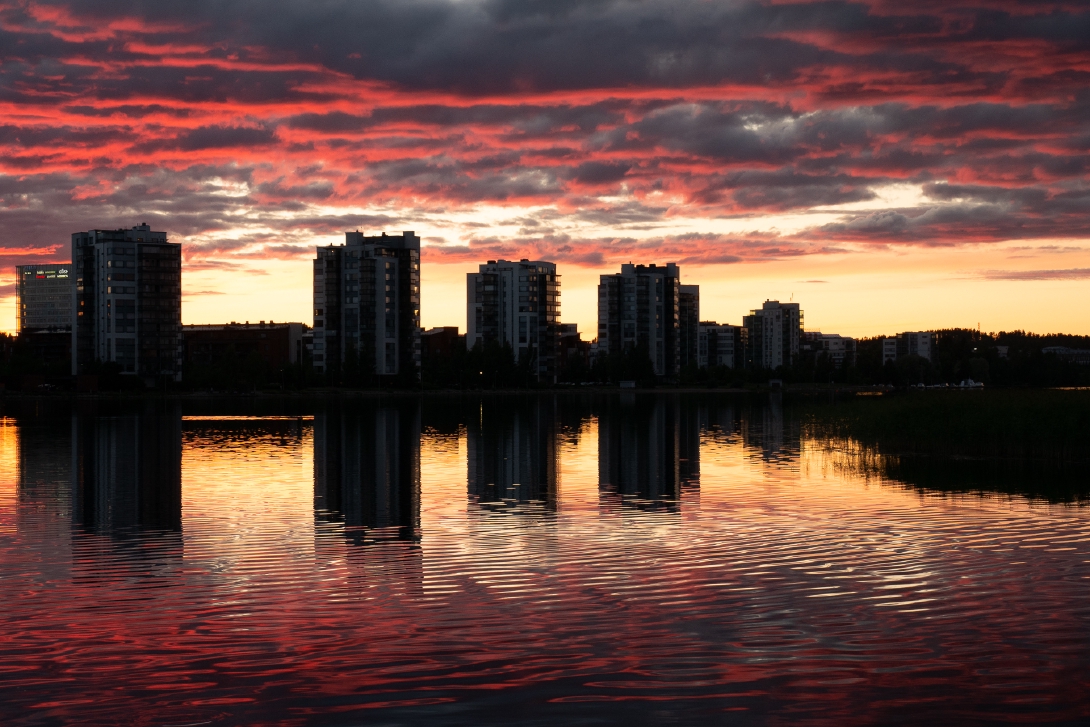 Kuva moderneista taloista järven rannassa punaisen auringonlaskun sävyttämänä.